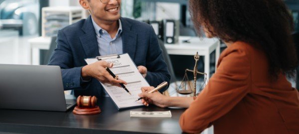 Mujer firmando un contrato mientras un abogado de seguros señala el documento en una oficina con mazo y balanza.
